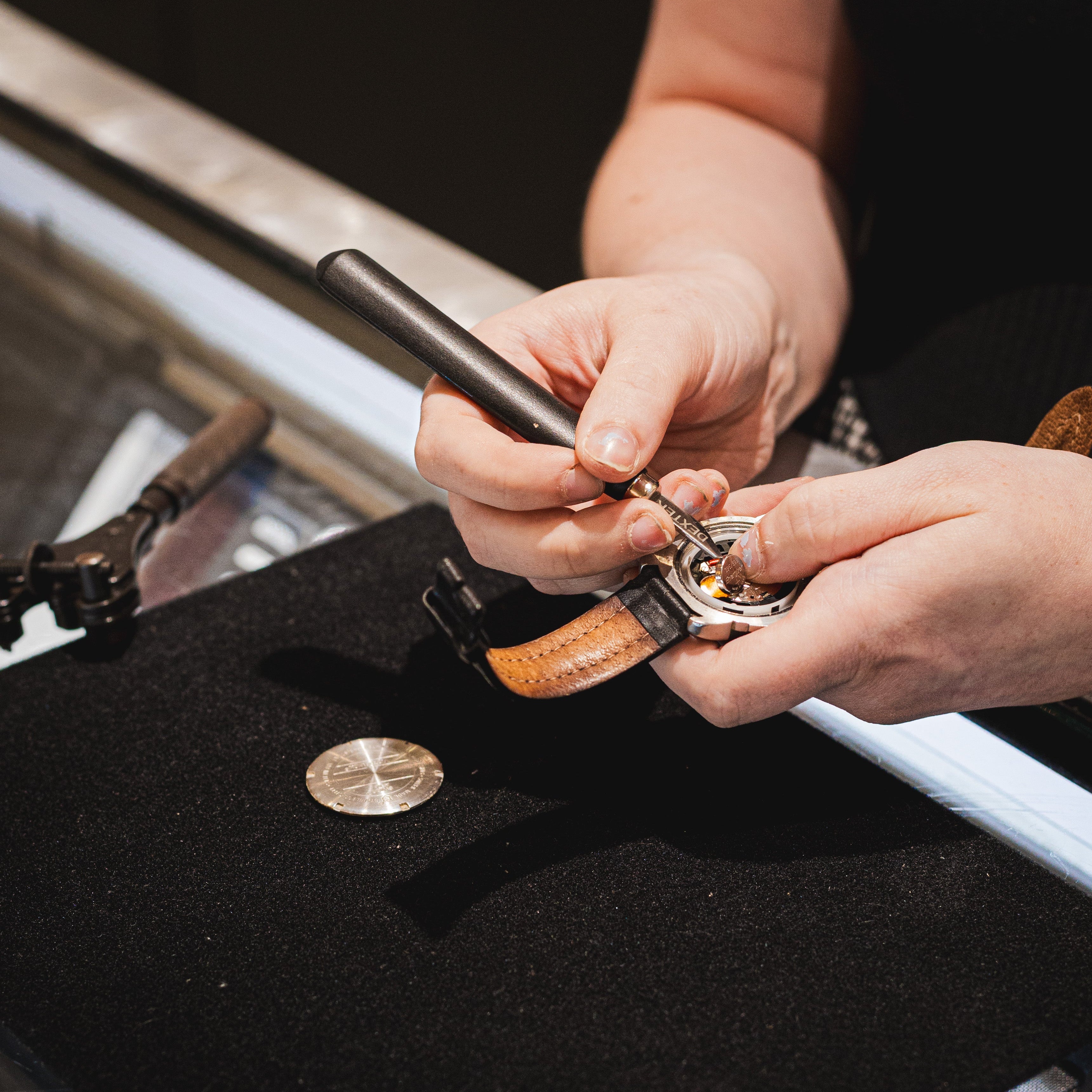 Close-up of a watchmaker's hands using a precision tool on a watch movement inside a watch case, with a black velvet work surface and watch parts in the background.