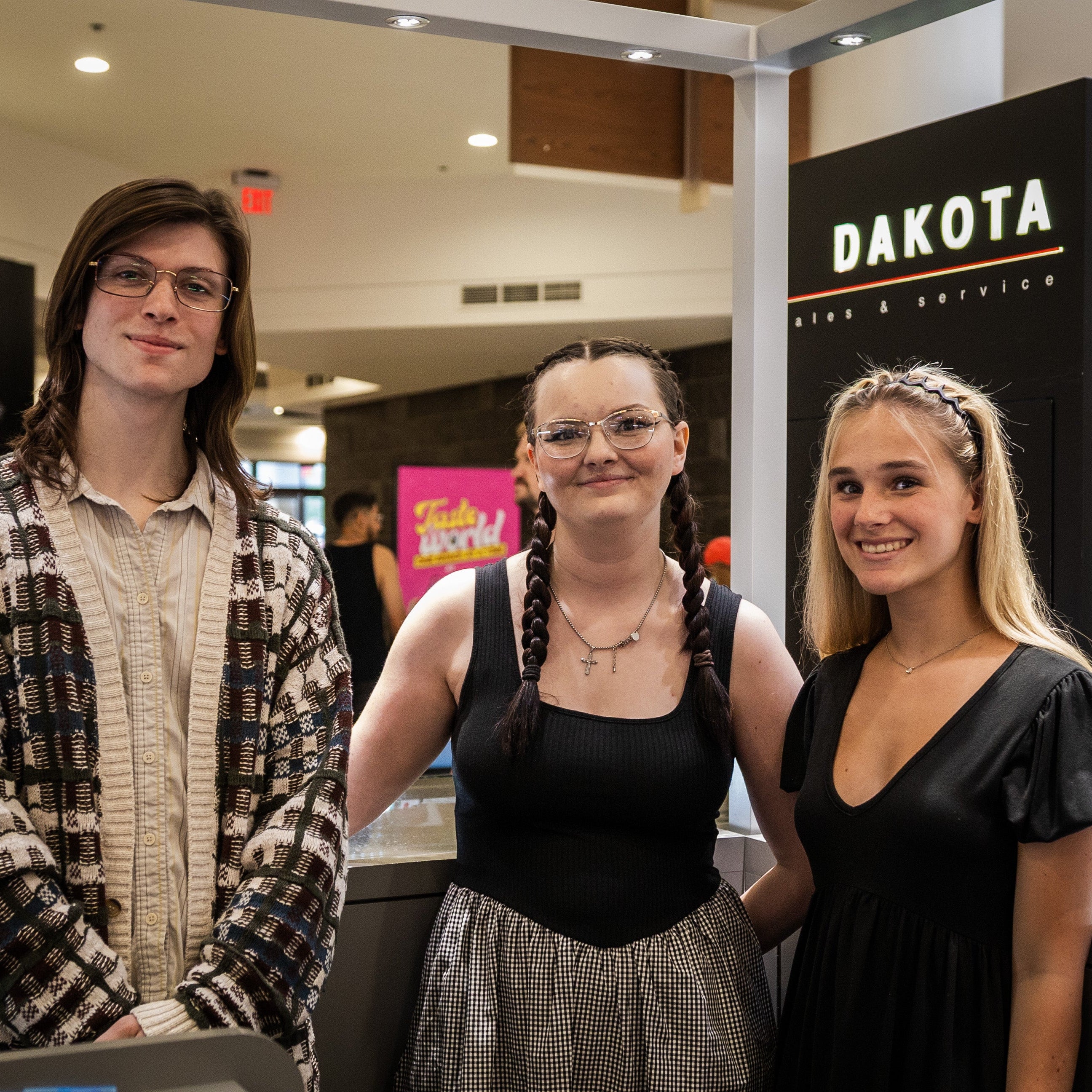 Three young people standing together in front of a DAKOTA signage structure inside a mall or event space. The person on the left wears glasses and a patterned button-up shirt, the center person wears glasses, a black sleeveless top, and a gray skirt, and the person on the right wears a black V-neck dress.