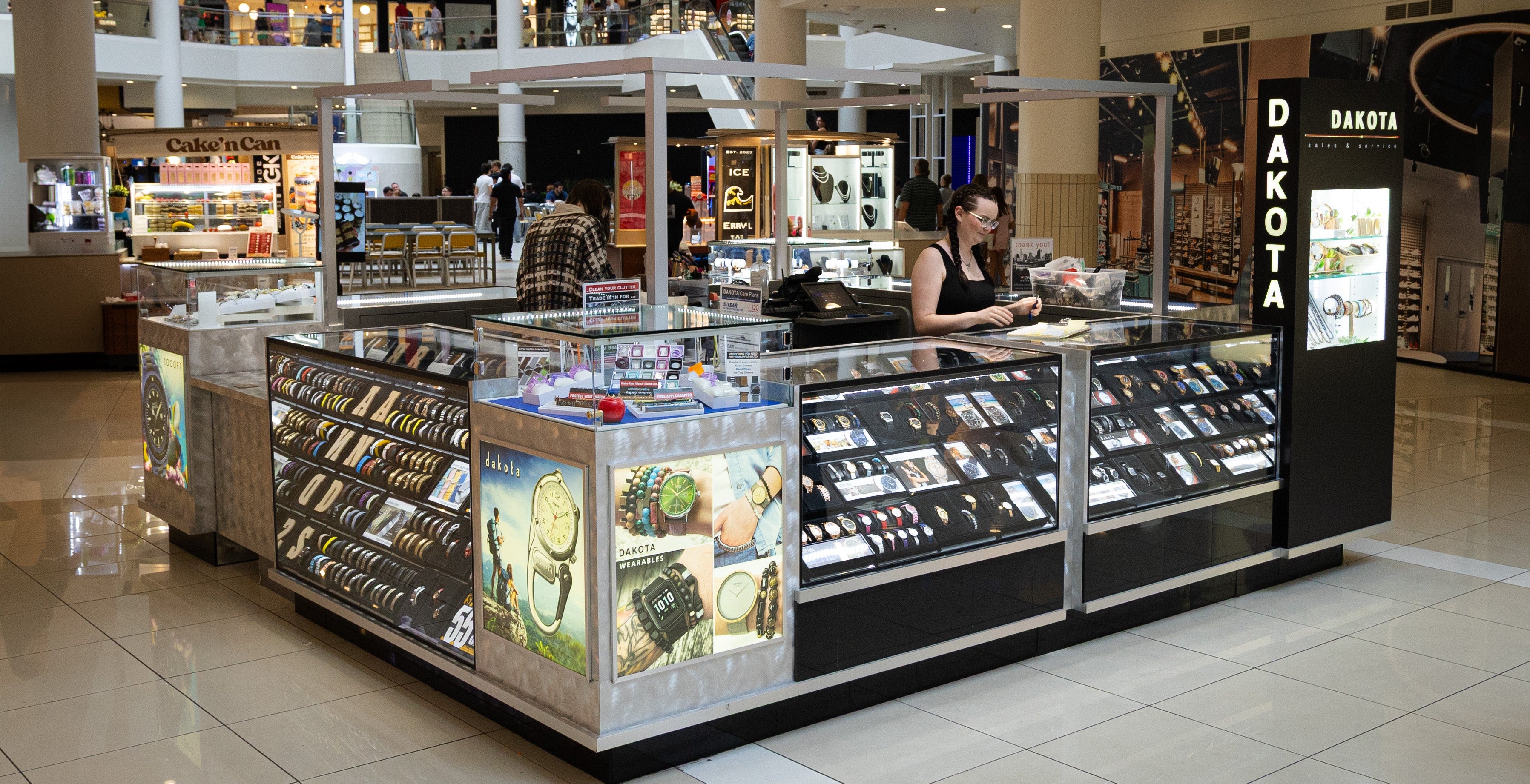 A retail display stand or kiosk in a shopping mall, featuring multiple glass display cases with jewelry and accessories, illuminated panels, and a vertical sign reading DAKOTA. A woman is seated behind the counter area, and the mall interior with tiled floors and upper levels is visible in the background.