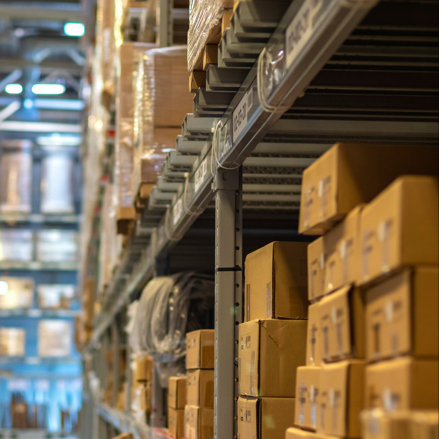 A blurred warehouse aisle with tall metal shelving stocked with brown cardboard boxes on the right side and more shelving on the left.