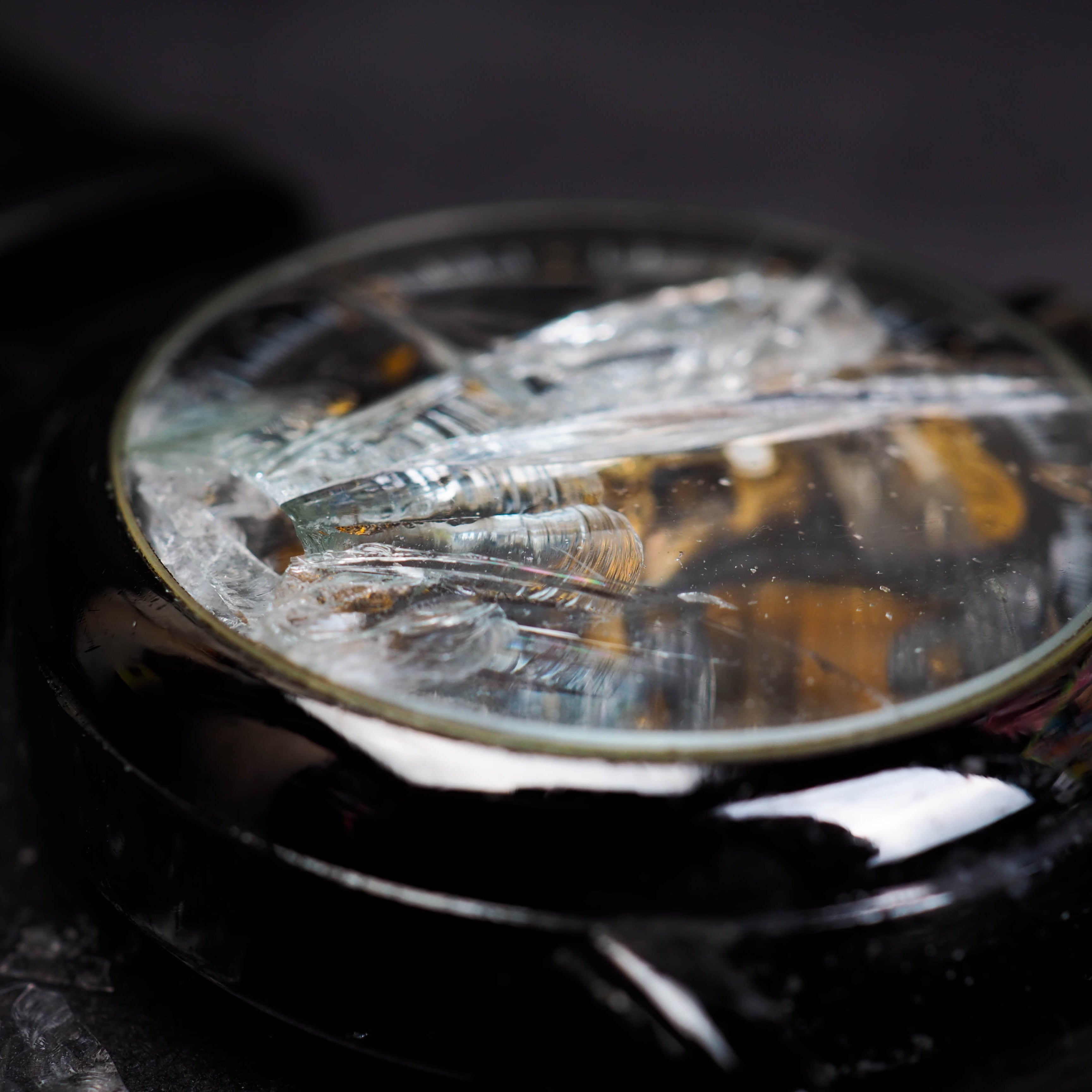 Close-up of a glossy black wristwatch with a severely shattered glass crystal, its jagged cracks catching iridescent light while revealing the gold mechanical movement beneath. Small glass fragments rest on a dark textured surface, with a matte black strap fading into a softly blurred background.
