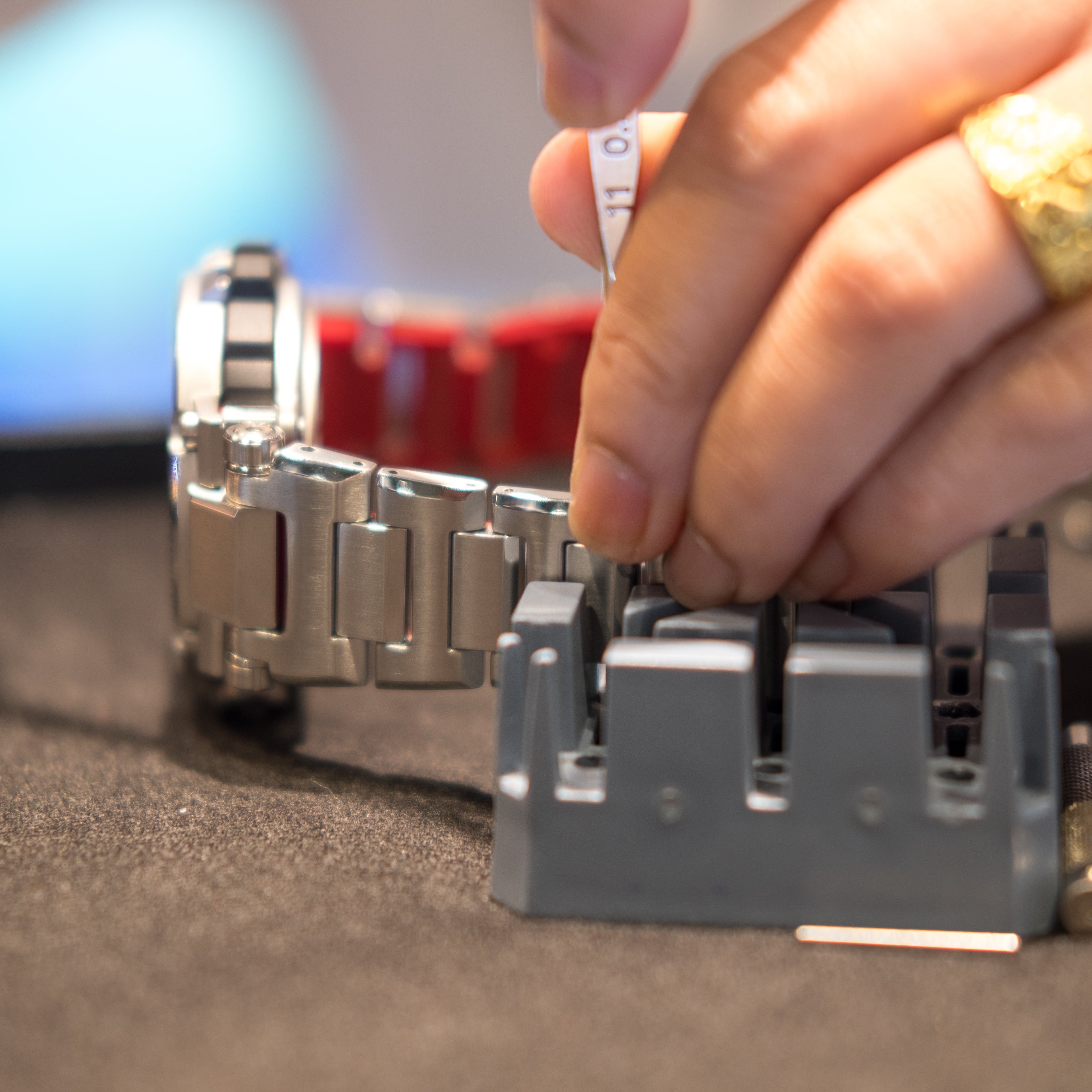 On a dark textured workbench, a chunky silver metallic-link watch with a red-lined strap rests beside a gray slotted holding block while a hand wearing a gold ring guides a white-handled pin punch into a link; loose metal pins and a knurled punch tool sit nearby, set against a soft blue bokeh background.