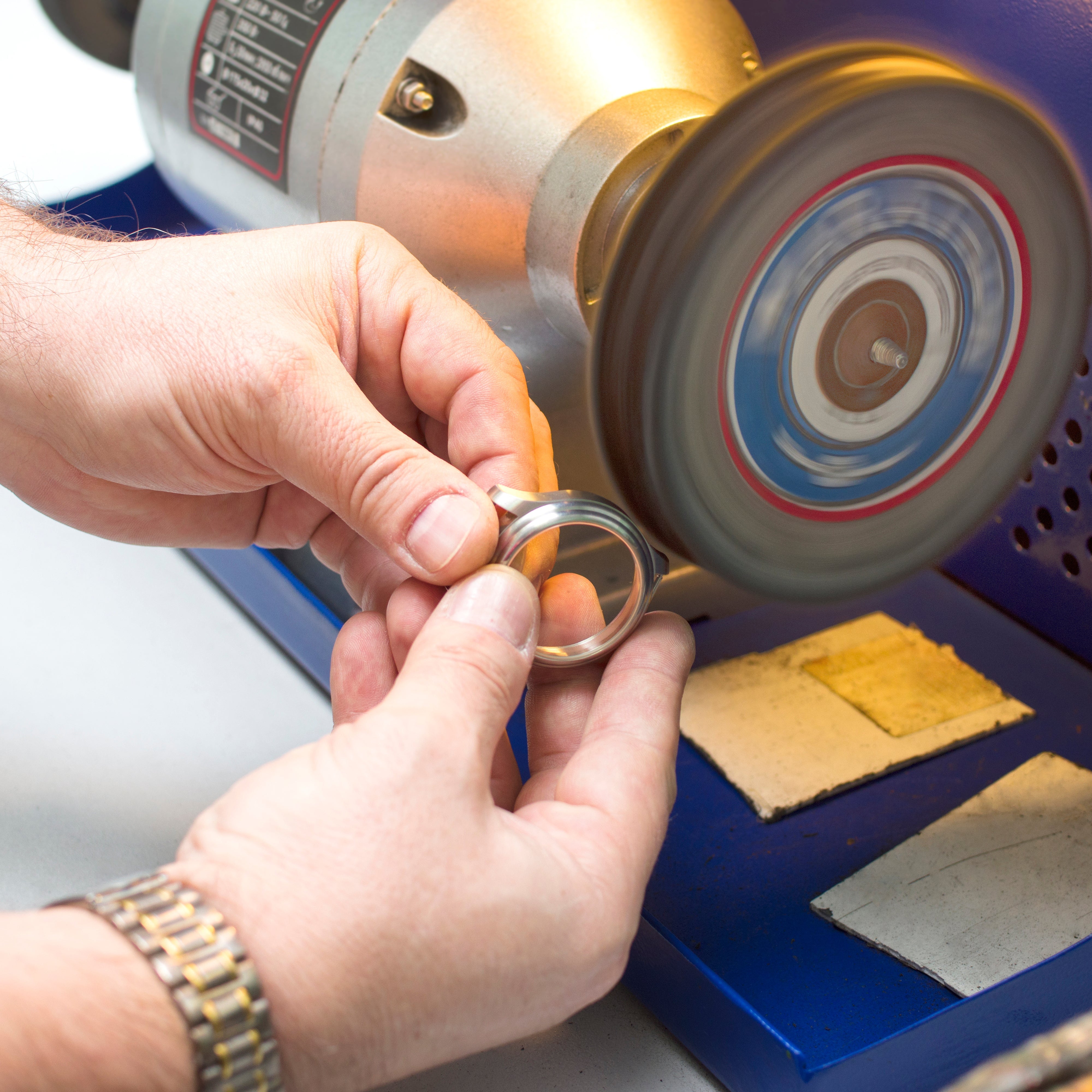 Two hands grip a polished silver watch bezel just inches from a rapidly spinning buffing wheel, its blue, white, and red concentric layers swirling into a soft motion blur. The wheel sits on a blue bench-mounted polishing machine with a gray motor housing, a ventilated base, and a technical spec label. One wrist wears a metallic watch band with silver links and warm gold accents, a quiet nod to the craft at hand. Bright, neutral workbench light frames the scene, highlighting the steady precision of finishi