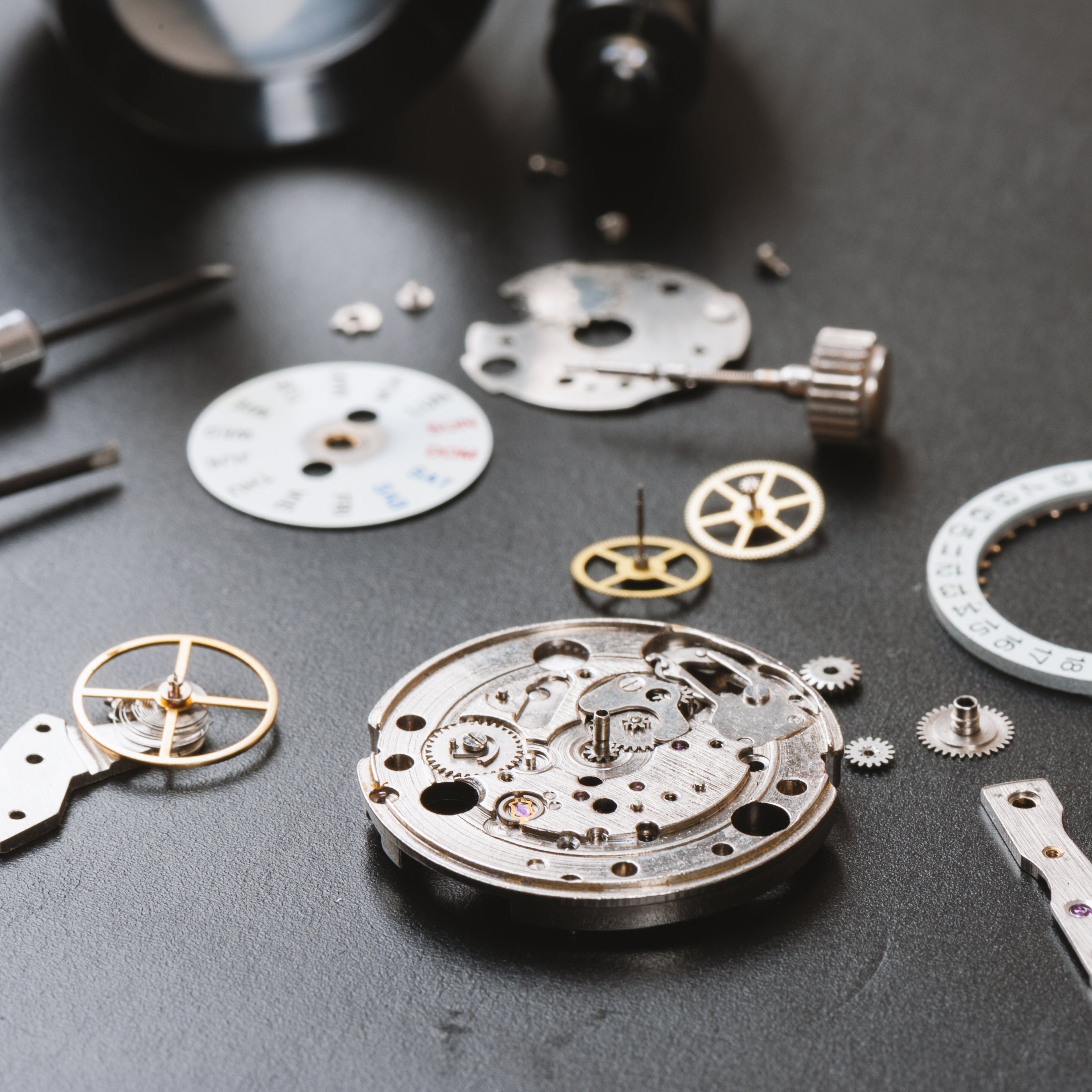 Flat-lay of a fully disassembled mechanical watch movement on a dark gray mat, showing the central brass and steel mainplate set with jewels and gears, a balance wheel with hairspring, a silver bridge fitted with purple ruby jewels, small brass gear wheels, a winding stem and crown, a white day disc reading SUN to SAT in blue and red, a white date ring numbered 1 to 31, scattered tiny screws, two knurled-handle precision screwdrivers on the left, and a black loupe at the top.