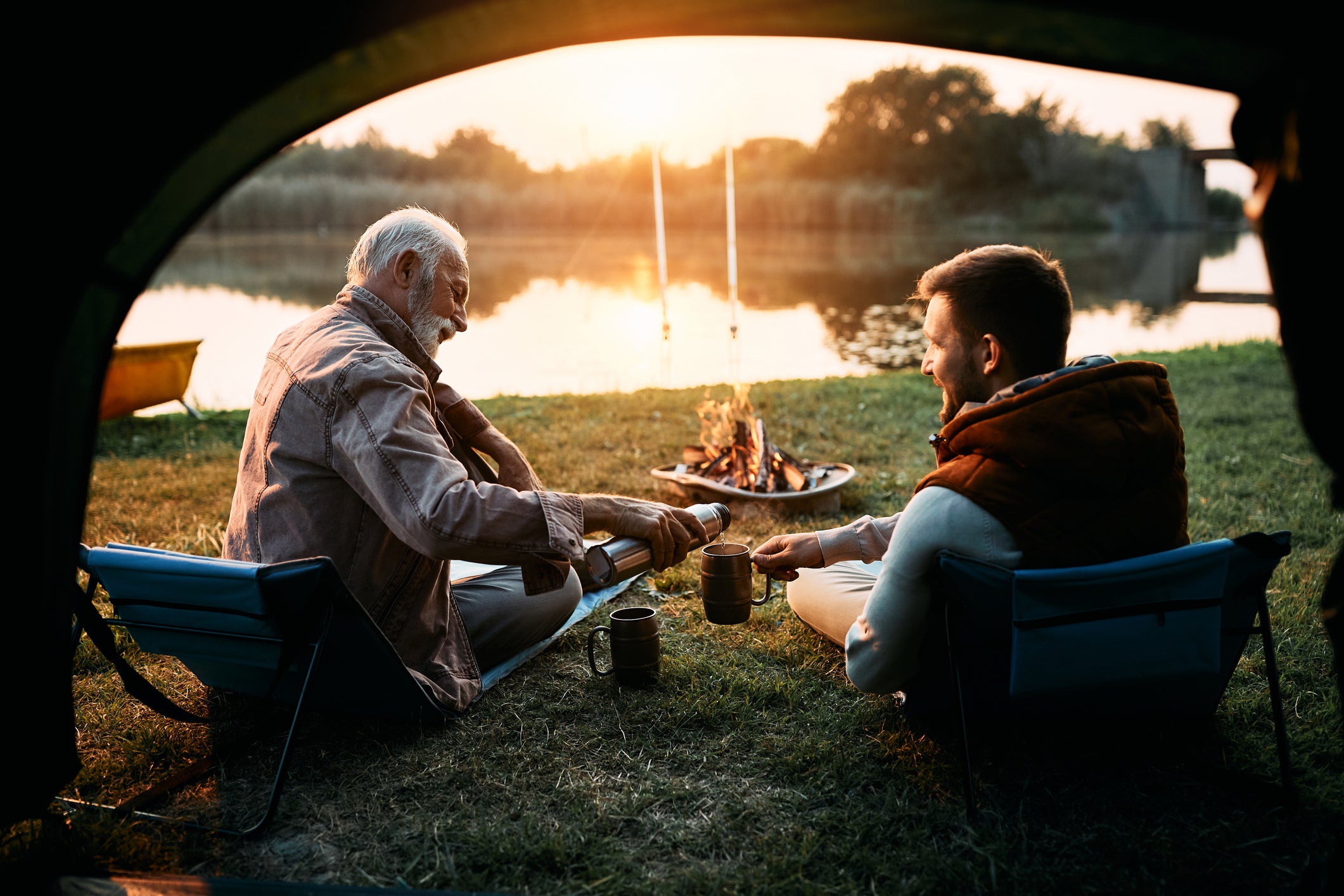 Senior fisherman and his son having a drink while camping by the water at sunset.