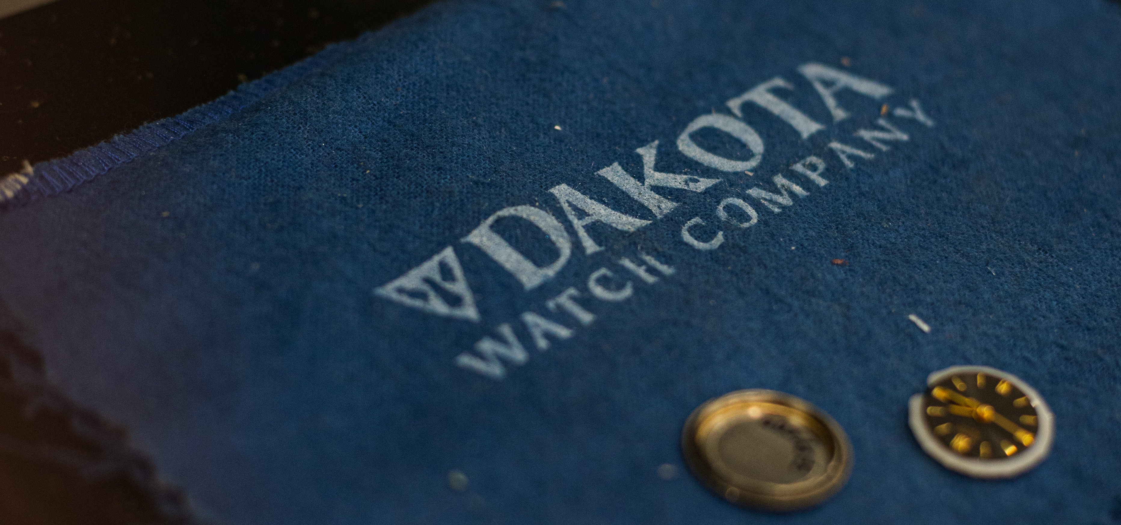 A gold-tone watch bracelet laid across a blue Dakota Watch Company microfiber cloth on a dark workbench, with two small watch parts placed on top.