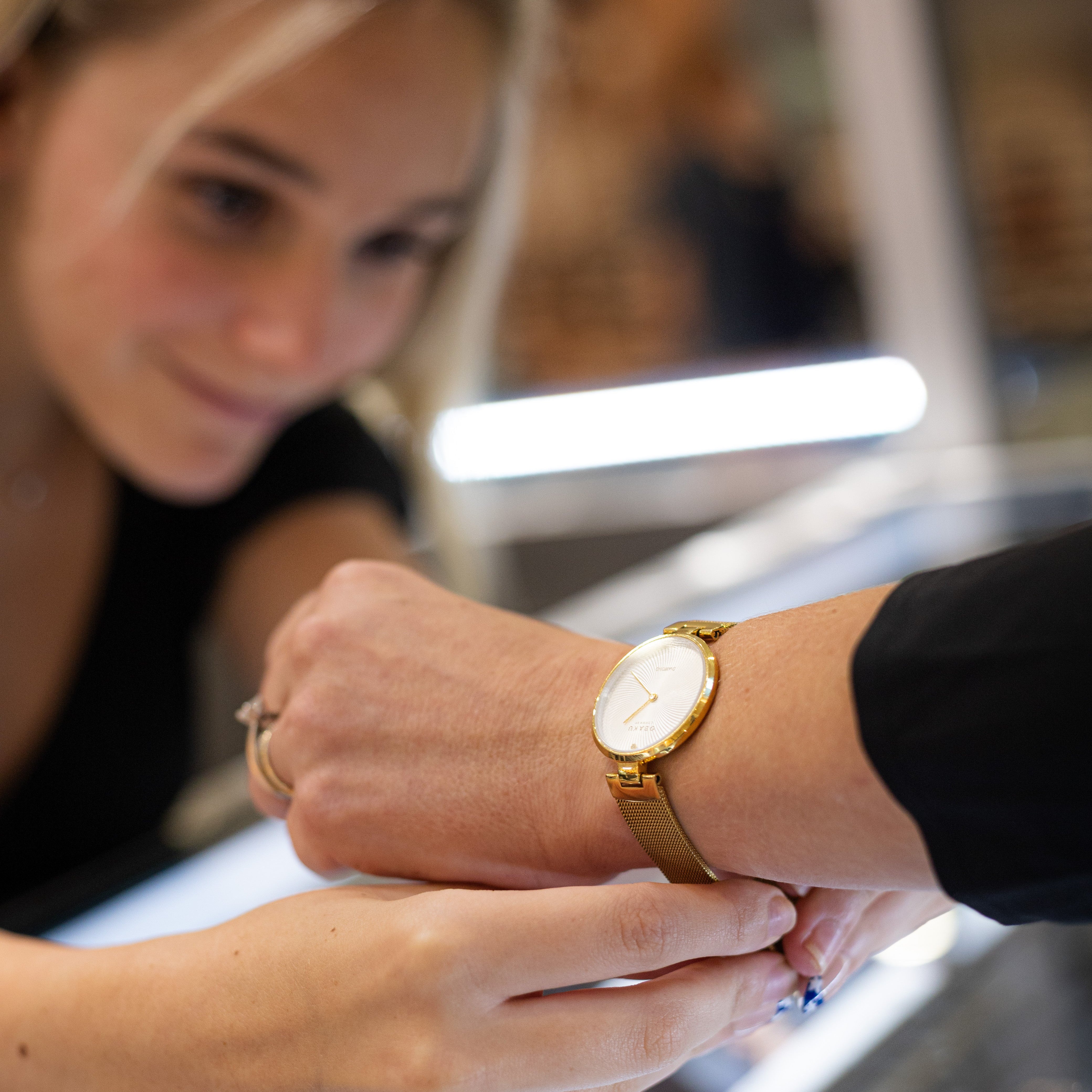 A close-up lifestyle shot of a woman wearing a gold-tone analog watch with a white dial, while another person adjusts or examines it near a jewelry display counter in a store.