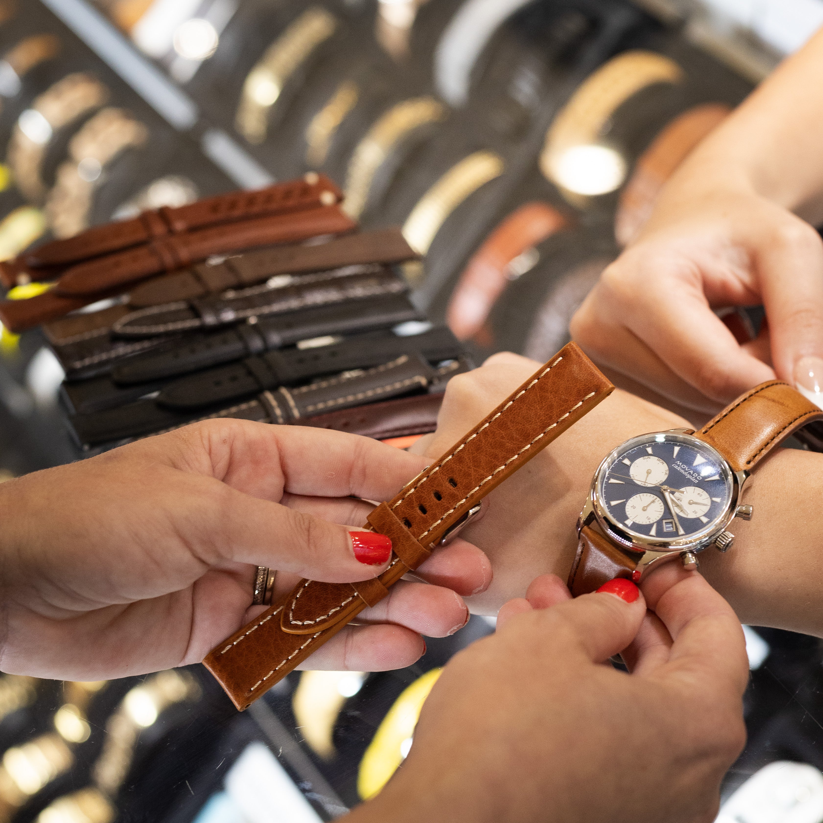 A close-up lifestyle shot of a watch being fitted with a brown leather strap by a watchmaker's hands. The watch has a silver-toned case and a dark dial with three sub-dials.