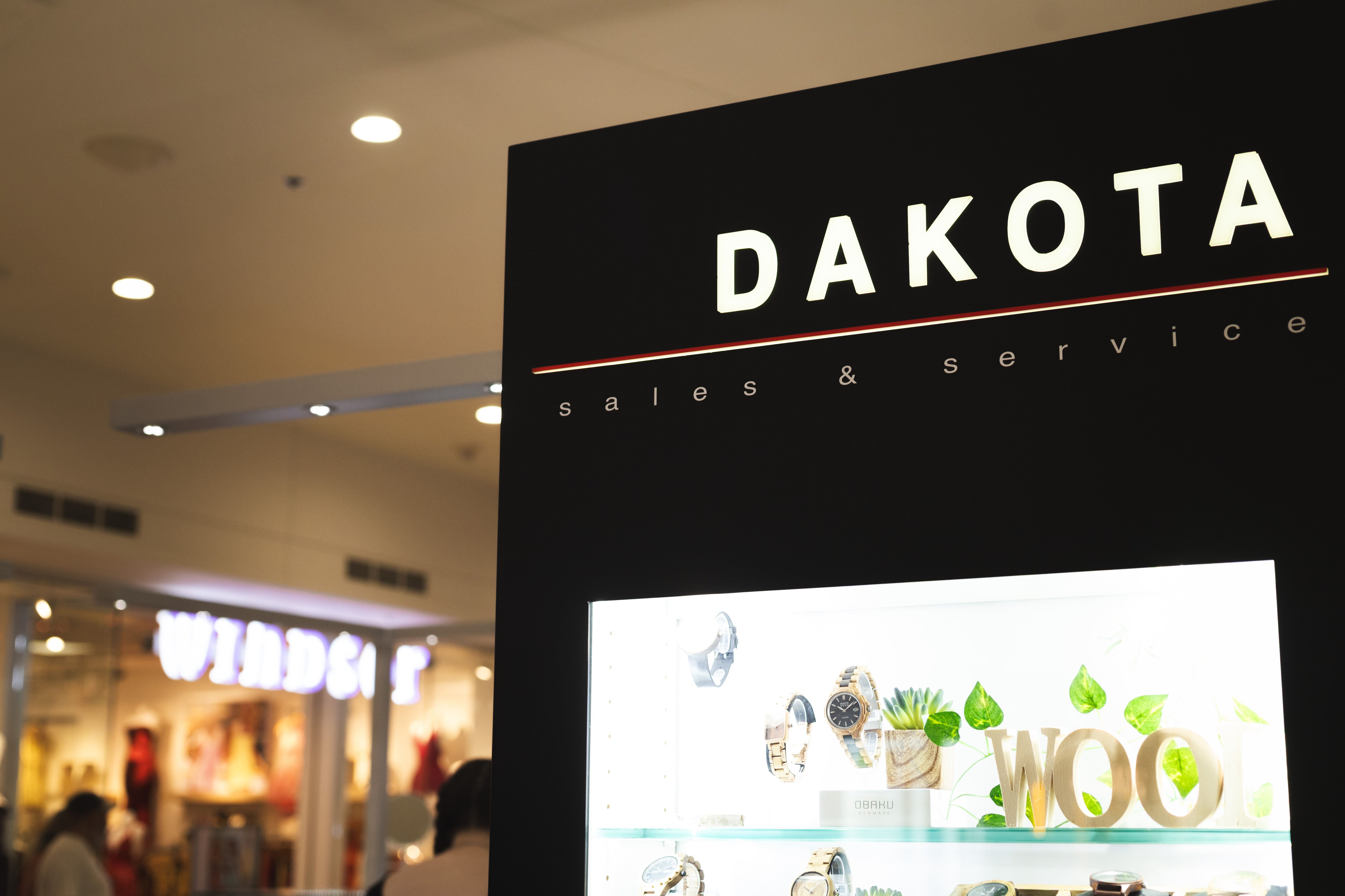 A large black retail display sign with the word 'DAKOTA' in white above a lit window display, photographed inside a shopping mall. Other store signage and blurred shoppers are visible in the background.